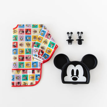 Bumkins Sleeved Bib: Mickey & Friends Check Mates is shown folded on a white background next to a Mickey-shaped plate and matching utensils.