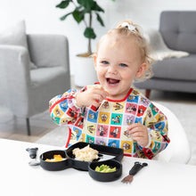 Load image into Gallery viewer, A toddler with a ponytail smiles while eating from a black plate on a white table, wearing Bumkins Sleeved Bib: Mickey &amp; Friends Check Mates.
