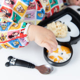 A child in Bumkins Sleeved Bib: Mickey & Friends Check Mates enjoys baby-led weaning from a Mickey-shaped plate.