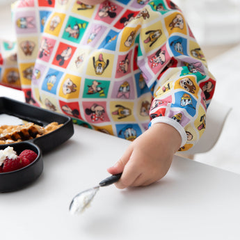 A child in Bumkins Sleeved Bib: Mickey & Friends Check Mates holds a spoon at a white table in front of a plate with raspberries.