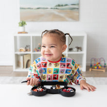 Load image into Gallery viewer, A toddler in Bumkins Sleeved Bib: Mickey &amp; Friends Check Mates smiles at a table with food, surrounded by shelves of toys in the background.
