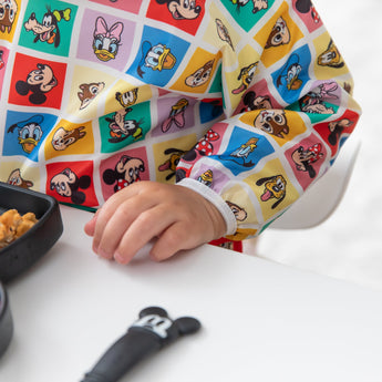 A child in Bumkins Sleeved Bib: Mickey & Friends Check Mates sits at a white table with a black dish and utensil.