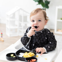 Load image into Gallery viewer, A boy in a high chair self feeds from a Mickey Mouse-shaped plate while wearing a Bumkins Sleeved Bib: Mickey Mouse Icon Black + White.
