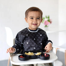 Load image into Gallery viewer, A toddler in a high chair, smiling with a divided plate of pasta and berries, wears a Bumkins Sleeved Bib: Mickey Mouse Icon Black + White.
