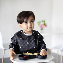 Load image into Gallery viewer, A dark-haired child in a high chair, wearing a Bumkins Sleeved Bib: Mickey Mouse Icon Black + White, eats from a black Mickey Mouse plate.
