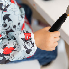 Load image into Gallery viewer, Close-up of a toddler in a Bumkins Sleeved Bib: Mickey Mouse holding a black-handled utensil.
