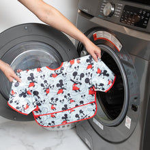 Load image into Gallery viewer, A person holds Bumkins Sleeved Bib: Mickey Mouse in front of an open front-loading washing machine.
