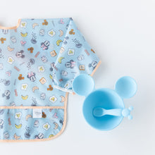 Load image into Gallery viewer, Bumkins Sleeved Bib: Mickey + Minnie Sunny Side Up, laid flat on a white background, is half visible next to a blue Mickey Mouse-shaped bowl and spoon set.
