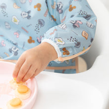 Load image into Gallery viewer, A child's hand reaches for food on a pink tray, wearing Bumkins Sleeved Bib: Mickey + Minnie Sunny Side Up.
