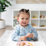 A child with pigtails smiles in a Bumkins Sleeved Bib: Mickey + Minnie Sunny Side Up in a high chair with snacks on a pink plate.