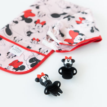 Bumkins Sleeved Bib: Minnie Mouse lays folded next to Minnie Mouse-shaped utensils on a white background.