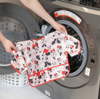 A person holds Bumkins Sleeved Bib: Minnie Mouse in front of an open front-loading washing machine.