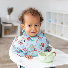 Load image into Gallery viewer, A curly-haired toddler in a high chair wears a Bumkins Sleeved Bib: Ariel with a green bowl and spoon on the tray.
