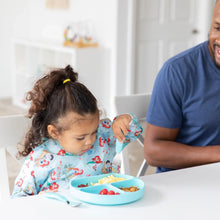 Load image into Gallery viewer, A child in a Bumkins Sleeved Bib: Ariel eats from a blue plate with a fork, sitting beside a smiling man at a white table.
