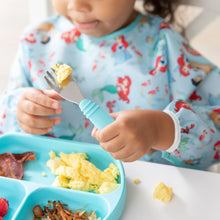 Load image into Gallery viewer, A child in a Bumkins Sleeved Bib: Ariel holds a fork with scrambled eggs by a blue sectioned breakfast plate at a white table.
