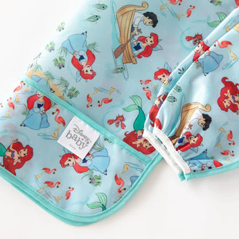 Close-up of Bumkins Sleeved Bib: Ariel folded on a white background, with focus on the front pocket and elastic cuff on the sleeves. 