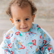 Load image into Gallery viewer, A toddler smiles at the camera, wearing a colorful Bumkins Sleeved Bib: Ariel with cartoon mermaids.
