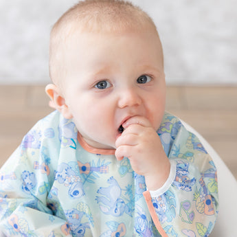 A baby in a Bumkins Sleeved Bib: Aloha Stitch sits curiously, hand in mouth, the waterproof fabric gleaming with colorful patterns.