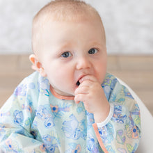 Load image into Gallery viewer, A baby in a Bumkins Sleeved Bib: Aloha Stitch sits curiously, hand in mouth, the waterproof fabric gleaming with colorful patterns.
