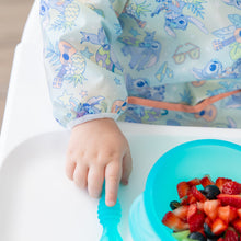 Load image into Gallery viewer, A child's hand wearing a Bumkins Sleeved Bib: Aloha Stitch with a blue spoon and a blue bowl of berries on a high chair tray.
