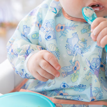 A baby enjoys self-feeding with a spoon, wearing a Bumkins Sleeved Bib: Aloha Stitch made of waterproof fabric.
