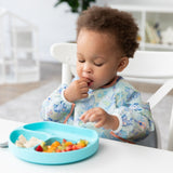 A toddler in a Bumkins Sleeved Bib: Aloha Stitch explores baby-led weaning, enjoying fruit and cheese from a divided blue plate on a white table.