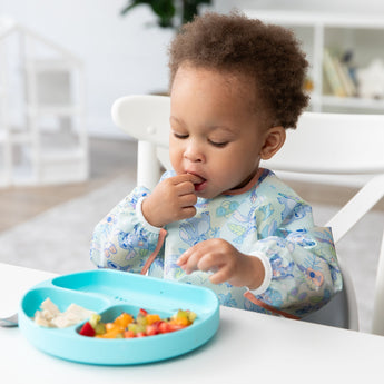 A toddler in a Bumkins Sleeved Bib: Aloha Stitch explores baby-led weaning, enjoying fruit and cheese from a divided blue plate on a white table.