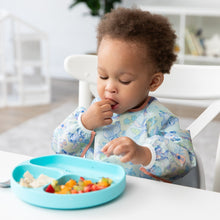 Load image into Gallery viewer, A toddler in a Bumkins Sleeved Bib: Aloha Stitch explores baby-led weaning, enjoying fruit and cheese from a divided blue plate on a white table.
