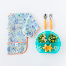 Load image into Gallery viewer, Bumkins Sleeved Bib: Aloha Stitch lays folded next to a blue divided plate with food and orange utensils on a white background.
