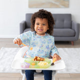 A young child smiling in a high chair wearing a Bumkins Sleeved Bib: Aloha Stitch, holding a spoon over food, with a sofa in the background.