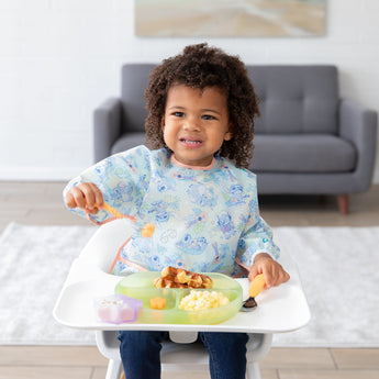 A young child smiling in a high chair wearing a Bumkins Sleeved Bib: Aloha Stitch, holding a spoon over food, with a sofa in the background.