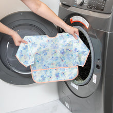Load image into Gallery viewer, A person holds a Bumkins Sleeved Bib: Aloha Stitch in front of a front-loading washing machine.
