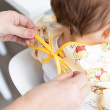 Load image into Gallery viewer, Close-up of adult hands tying the ribbon closure of a Bumkins Sleeved Bib: Winnie Loves Hunny around a child's neck.
