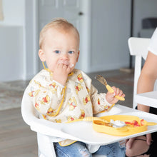 Load image into Gallery viewer, A baby in a high chair, wearing a Bumkins Sleeved Bib: Winnie Loves Hunny, enjoys baby-led weaning with a yellow fork and plate.

