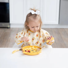 Load image into Gallery viewer, A toddler in a Bumkins Sleeved Bib: Winnie Loves Hunny uses a fork to eat from a yellow plate, exploring baby-led weaning at a white table.
