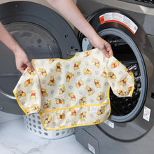 Load image into Gallery viewer, A person holds Bumkins Sleeved Bib: Winnie Loves Hunny in front of an open front-loading washing machine.
