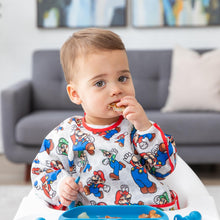 Load image into Gallery viewer, A boy in a high chair eats curiously from a blue plate in a living room while wearing a Bumkins Sleeved Bib: Super Mario™ Classic.
