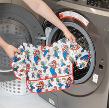 Load image into Gallery viewer, A person holds Bumkins Sleeved Bib: Super Mario™ Classic in front of an open front-loading washing machine.
