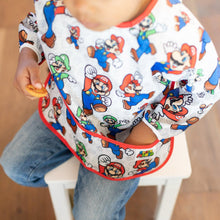 Load image into Gallery viewer, A child in a Bumkins Sleeved Bib: Super Mario™ Classic sits on a white chair with their hand in the pocket.
