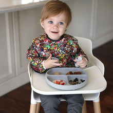 Load image into Gallery viewer, A toddler in a Bumkins Sleeved Bib: Super Mario™ Mashup sits in a high chair with snacks in a gray divided plate.
