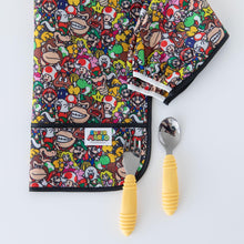 Load image into Gallery viewer, Bumkins Sleeved Bib: Super Mario™ Mashup lays folded near a yellow-handled spoon &amp; fork on a white background.
