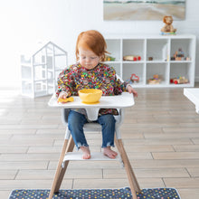 Load image into Gallery viewer, In a bright playroom, a child sits in a high chair, wearing a Bumkins Sleeved Bib: Super Mario™ Mashup and eating from a yellow bowl.

