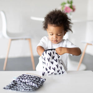 A curly haired toddler opens a Bumkins Reusable Snack Bag, Large 2-Pack: XOXO & Hearts at a white table as the other bag sits nearby.