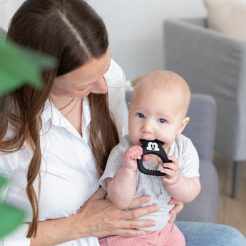 A woman holds a baby chewing a Bumkins Silicone Flat Teether: Mickey Mouse made from LFGB-grade silicone.