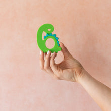 Load image into Gallery viewer, A hand holds Bumkins Silicone Teether: Dinosaur in front of a textured light orange wall.
