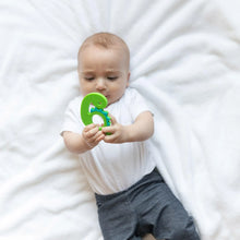 Load image into Gallery viewer, A baby in white outfit on a white blanket holds a Bumkins Silicone Teether: Dinosaur.
