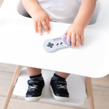 Load image into Gallery viewer, A toddler grips a Bumkins Silicone Teether: Nintendo SNES™ Controller in a high chair with black sneakers and white socks visible.
