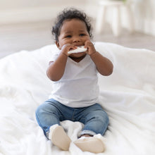 Load image into Gallery viewer, A baby in a white shirt and blue jeans happily chews on a Bumkins Silicone Teether: Unicorn while sitting on a soft blanket.
