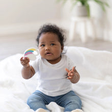 Load image into Gallery viewer, A baby on a white blanket holds a Bumkins Silicone Teether: Unicorn with a plant blurred in the background.
