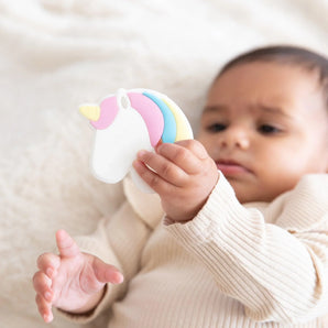 A baby in a beige outfit holds a Bumkins Silicone Teether: Unicorn on a soft, light-colored blanket.